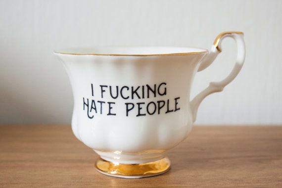 a white tea cup with gold trim is sitting on a wooden table. the cup has the phrase "i fucking hate people" written on it in black lettering. the background is a white wall. the tea cup appears to be a sarcastic or humorous decoration.