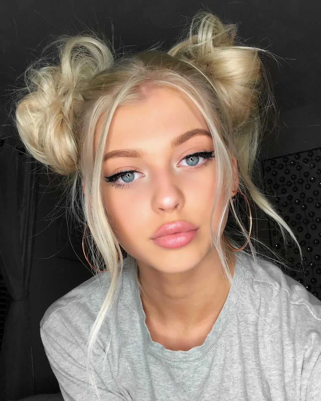 a closeup portrait of a young woman with blonde hair styled in space buns. she has blue eyes, wears eyeliner and lip gloss, and is wearing large hoop earrings and a gray tshirt.