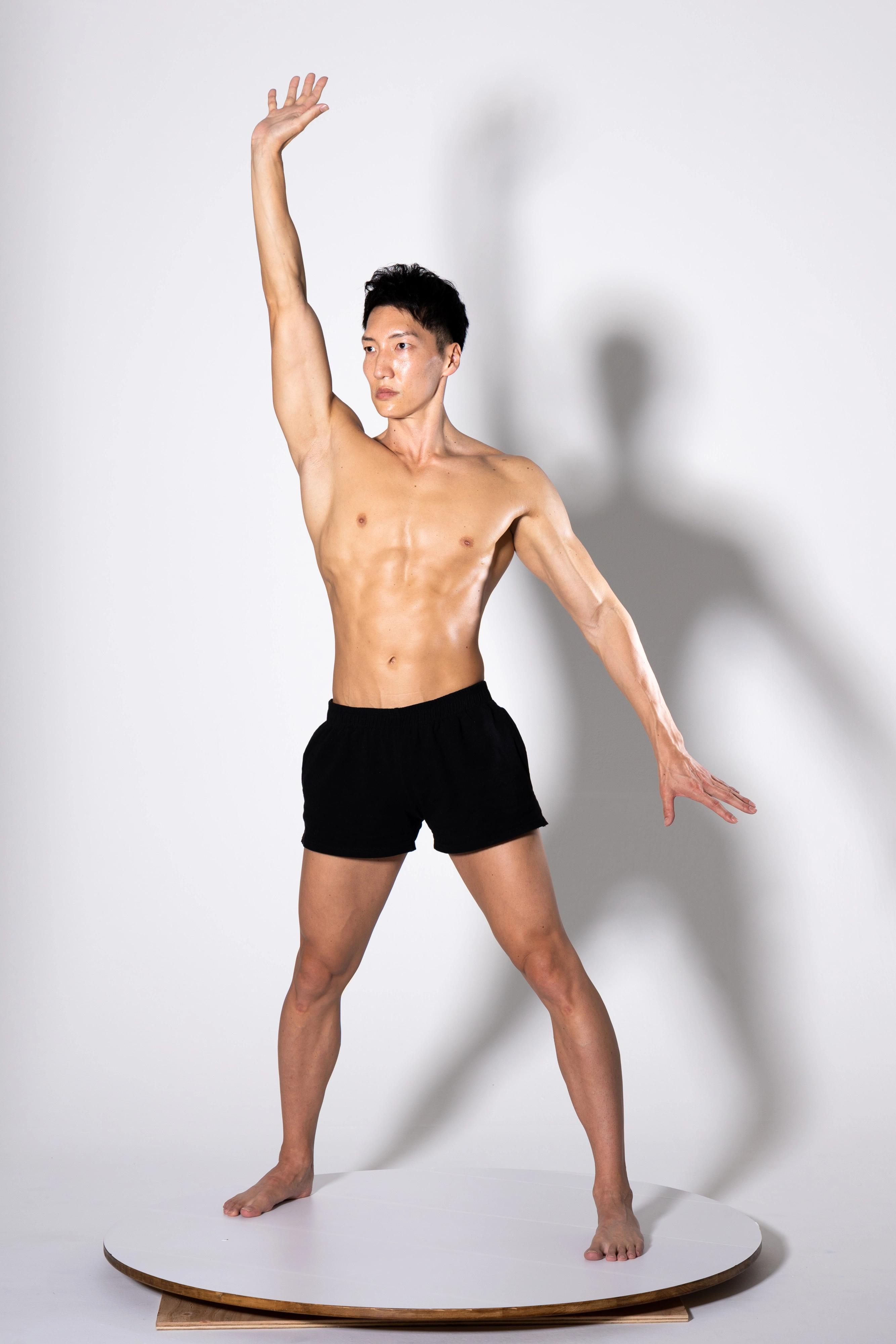 a muscular, athletic young man with a toned physique stands on a white turntable against a white background. he is shirtless, wearing only black shorts, and has one arm raised overhead. his pose suggests strength, flexibility, and athleticism. the image is welllit and shows a clear view of his body.  the lighting and background create a clean, professional look.