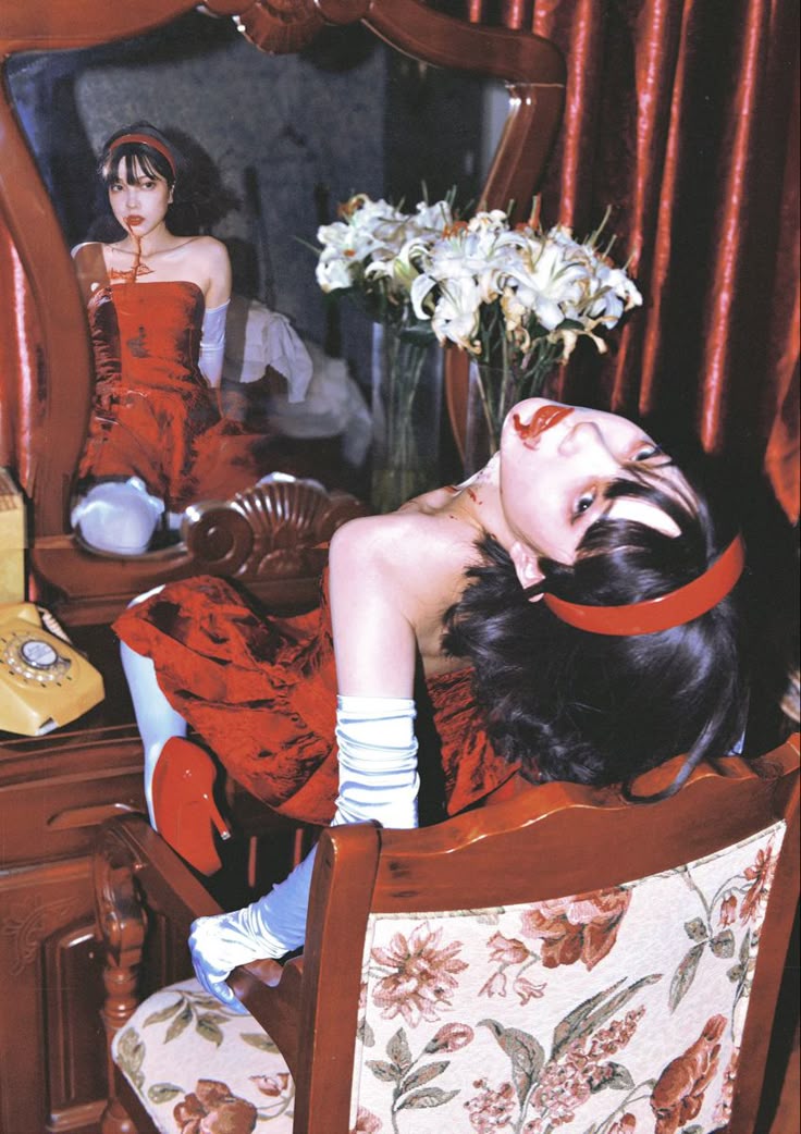a woman in a red dress and white gloves is slumped in a chair, with blood smeared around her mouth and neck. her head is tilted back, and she has a red headband in her dark hair. she is positioned in front of a vanity with a mirror, in which her reflection can be seen. the vanity also holds a vase of white lilies and an oldfashioned telephone. the chair is upholstered with a floral pattern, and a red curtain hangs in the background. the overall image has a vintage and somewhat unsettling aesthetic.
