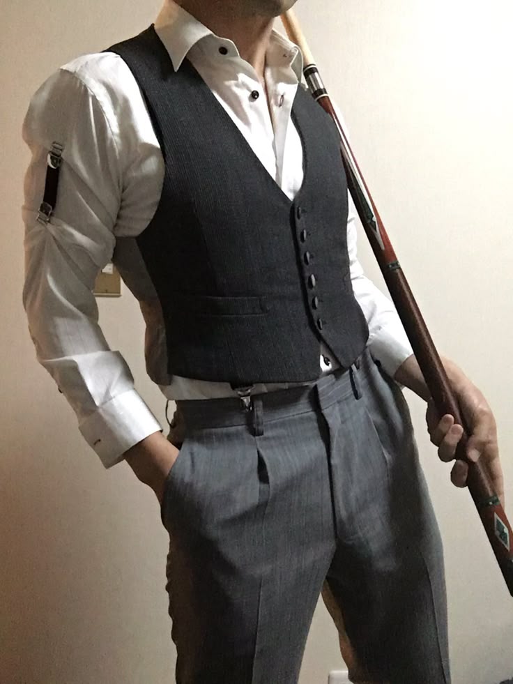 a man stands indoors wearing a white buttonup shirt with arm garters, a gray vest, and gray pleated pants with suspenders. he has one hand in his pocket and is holding a billiards cue.