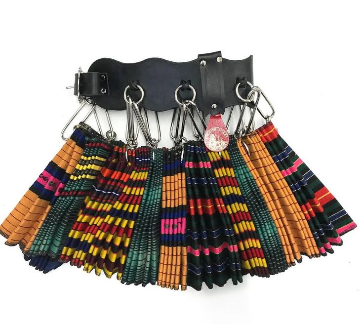 a black leather belt with multiple metal rings attached to it hangs from a white wall. attached to each ring is a folded fabric swatch with colorful african print patterns, resembling a skirt or decorative piece.