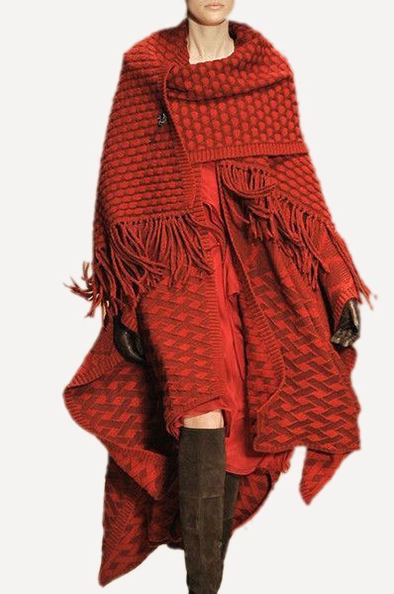 a woman wearing a red knitted cape with fringes on the bottom edge. the cape is made of a thick knit and has a loose fit. the model is also wearing long boots that match the color of the cape. she looks elegant and stylish in this outfit.