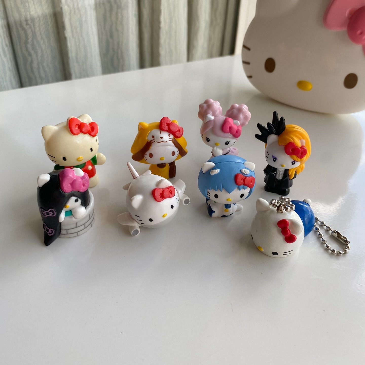 the image shows a collection of hello kitty figurines and a keychain on a white surface. there are eight figurines visible, each with a different design. one of the figurines is a keychain. in the background, there is a larger hello kitty object.