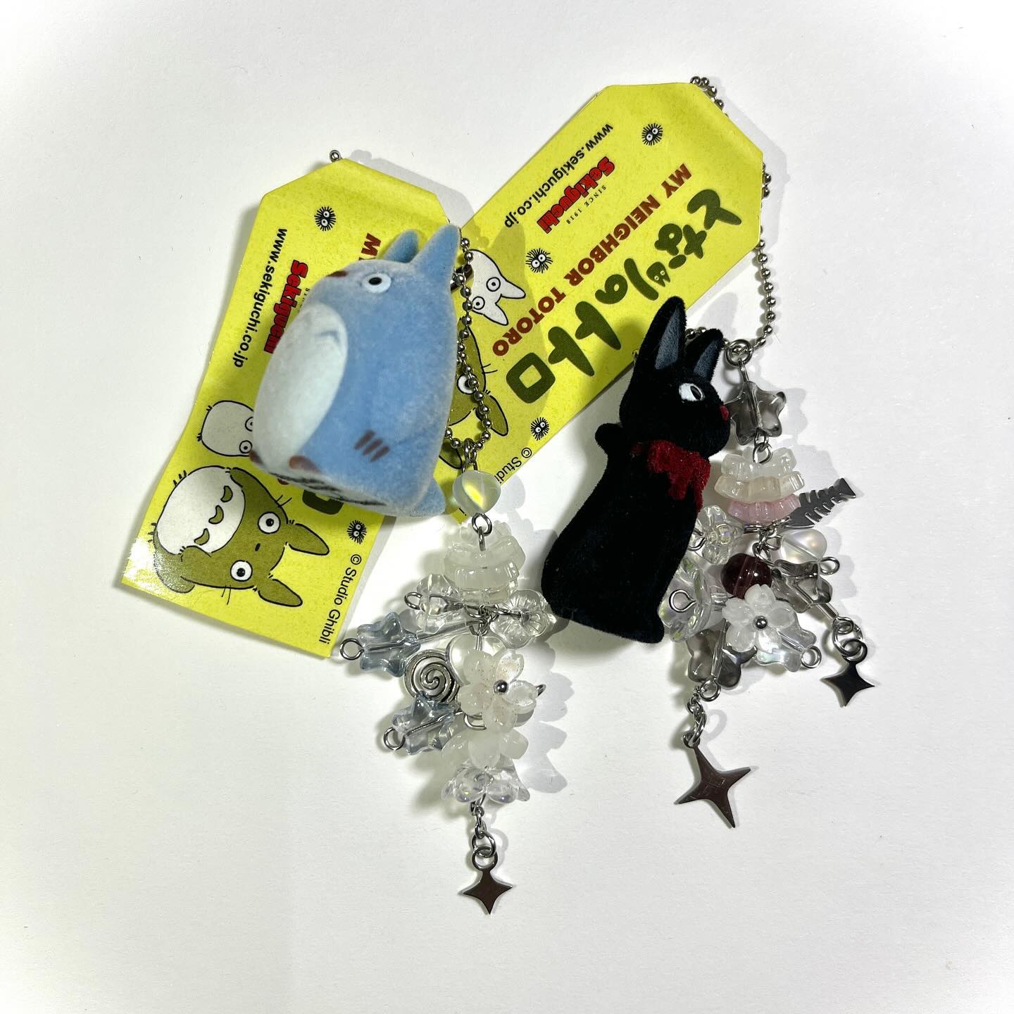 two studio ghibli "my neighbor totoro" keychains with yellow sekiguchi tags attached, featuring a blue totoro plush and a black soot sprite plush, both adorned with decorative beaded dangles.