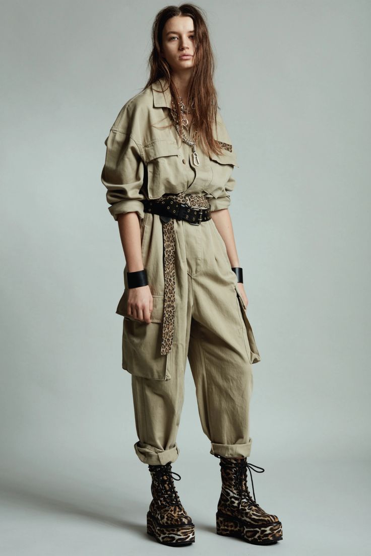 a young woman model in a stylish fashion photoshoot. she's wearing a tan, utilitystyle jumpsuit with a leopard print belt. she's also wearing a silver necklace with a pendant, black bracelets on both wrists, and brown, leopard print boots. her hair is long and brown and she's looking directly at the camera.  the background is a simple white wall.