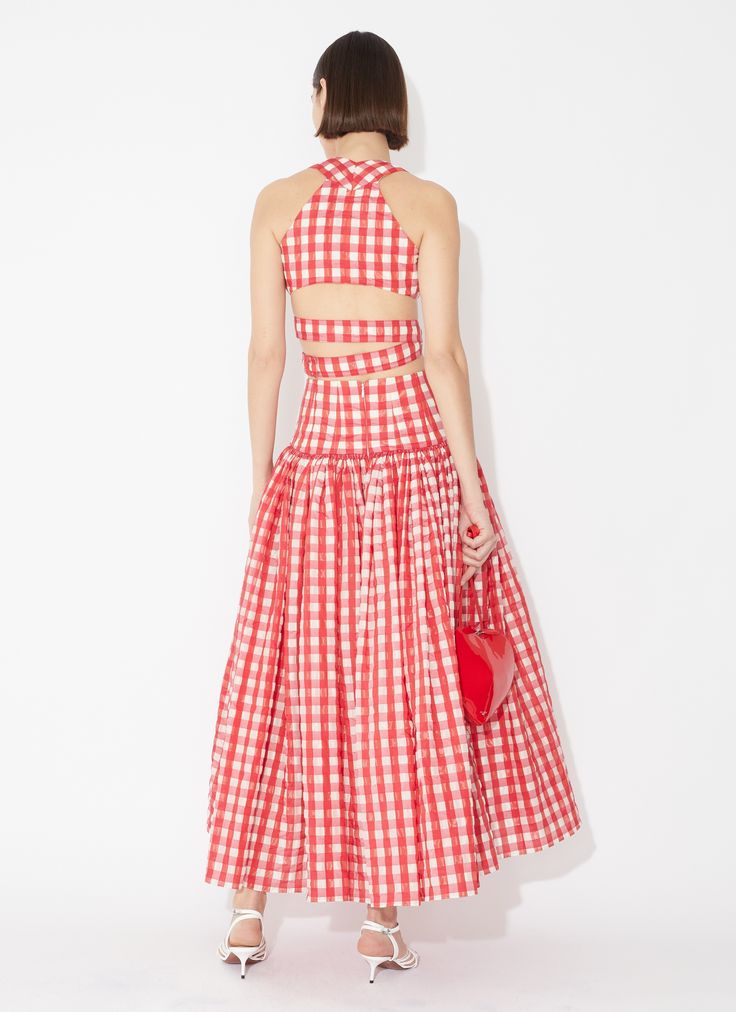 a woman is standing with her back to the camera, wearing a red and white checkered dress. the dress consists of a crop top with an exposed back and a full, flowing skirt. she is holding a red, heartshaped purse in her left hand. she is wearing white highheeled sandals. the background is a plain white.
