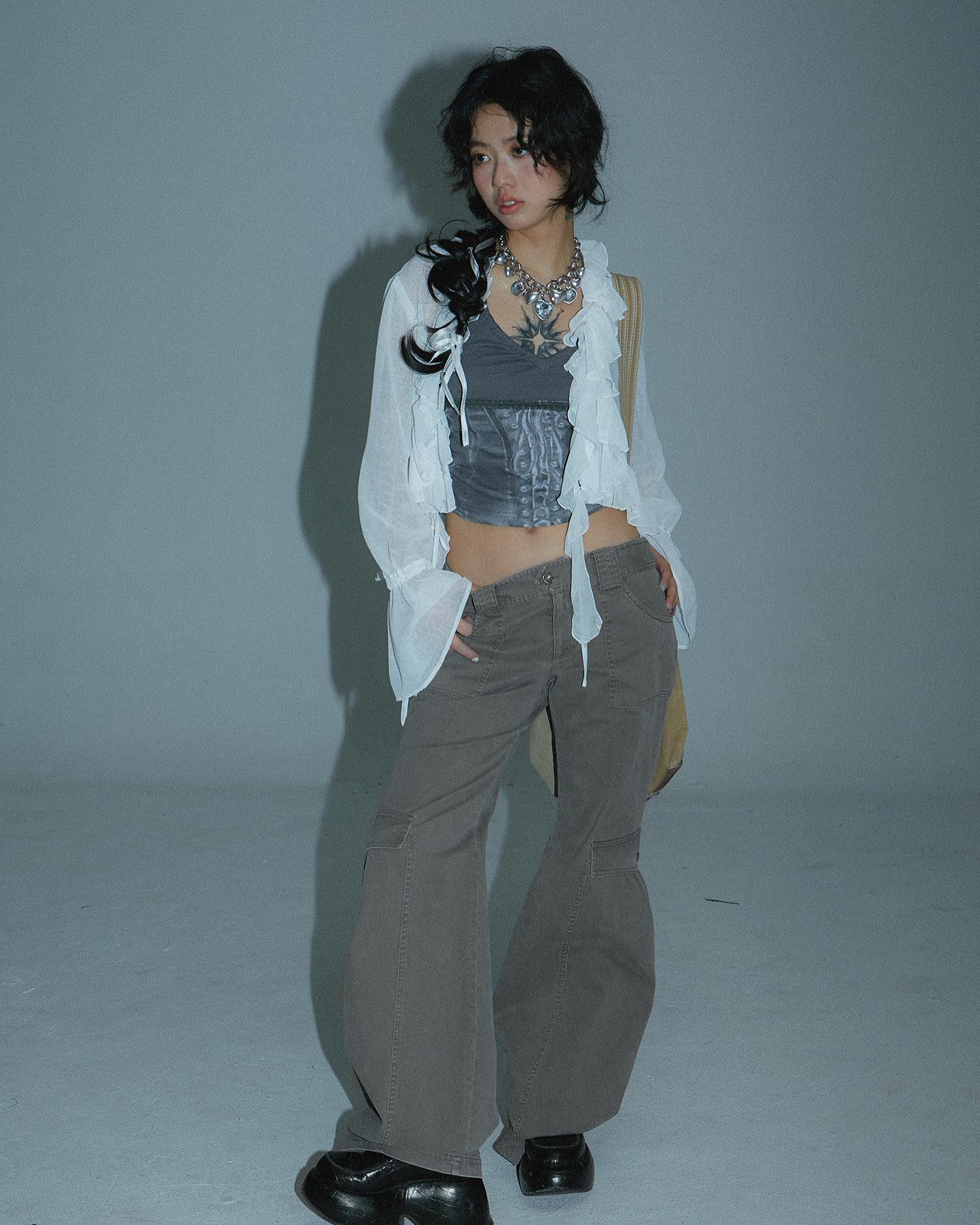 a fullbody shot of a young woman posing in a studio. she is wearing loosefitting, grey wideleg cargo pants, and an offwhite, sheer, ruffled blouse with a grey corset top underneath. the outfit is accessorized with a silver necklace. her dark hair is styled loosely. the overall aesthetic leans toward grunge or alt fashion with a hint of y2k/90s influence.  the backdrop is a simple, light grey. she's wearing black shoes and is standing with a slightly angled pose.