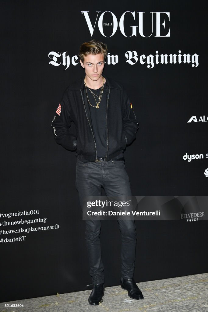 a full shot of a male model posing in front of a vogue italia backdrop at an event. he is wearing a black jacket, black tshirt, black jeans, and black boots. he has a gold chain around his neck. the backdrop has the vogue italia logo and the words "the new beginning" written on it. there are also logos for dyson and belvedere vodka. a getty images watermark and credit is visible.