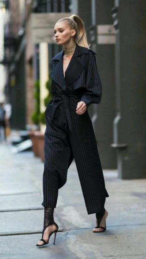 a woman in a black and white pinstripe jumpsuit, blazer, and heels walks confidently down a city street. she is accessorized with a gold chain necklace and a black handbag. her hair is pulled back in a sleek ponytail. the photo captures her effortlessly chic style.