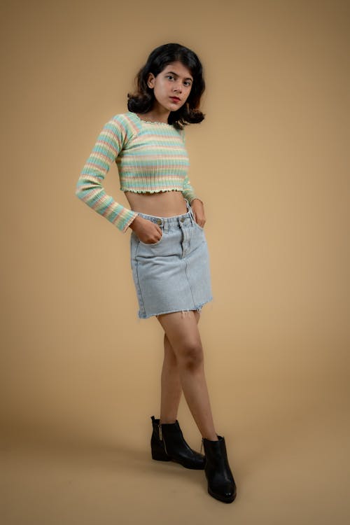 a young adult woman is posing in a studio shot. she is wearing a striped crop top and a denim skirt, paired with boots. she has a neutral expression and is looking slightly to the side.