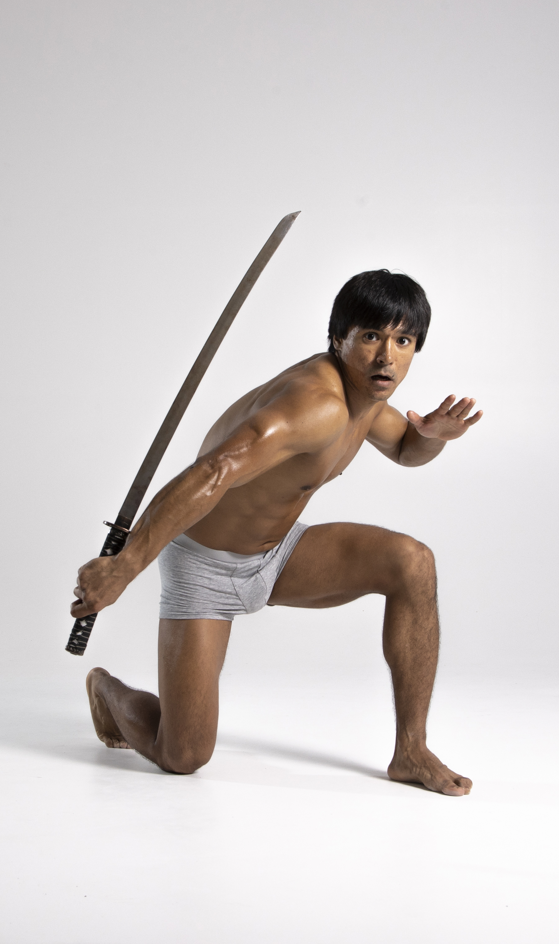a shirtless asian man is kneeling on one knee while holding a katana sword. he is wearing gray underwear and has a defensive posture. the background is a plain white color.