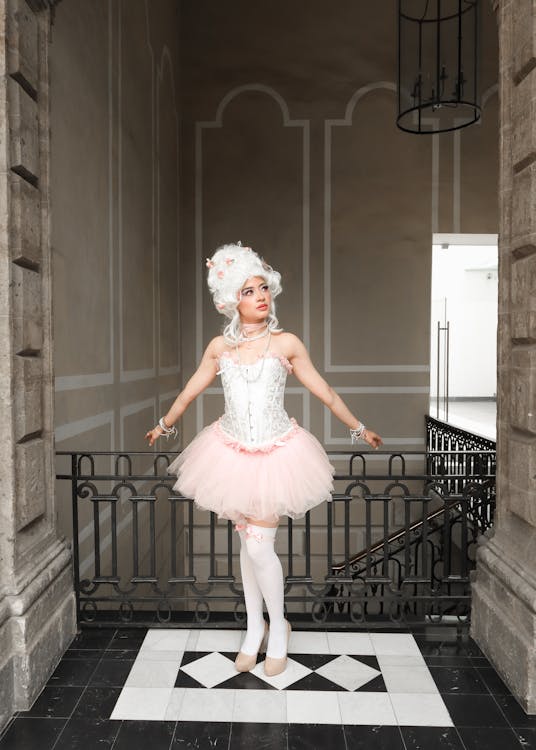 a fulllength portrait of a young woman dressed in a rococostyle costume. she is wearing a white corset, a pink tutu, white stockings, and heels. her wig is styled in a large, elaborate updo typical of the era.  the woman is standing on a staircase in a grand building, with a black wroughtiron railing visible. the overall aesthetic is elegant and vintage. she is looking upwards towards an unseen point.  the background features grand architecture with a muted color scheme, complementing the costume.