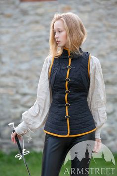 a blonde woman, dressed in a black quilted vest with yellow trim and black pants, stands holding a sword. she is outdoors against a backdrop of a weathered stone wall. the photo is taken at eye level and illuminated by natural light.