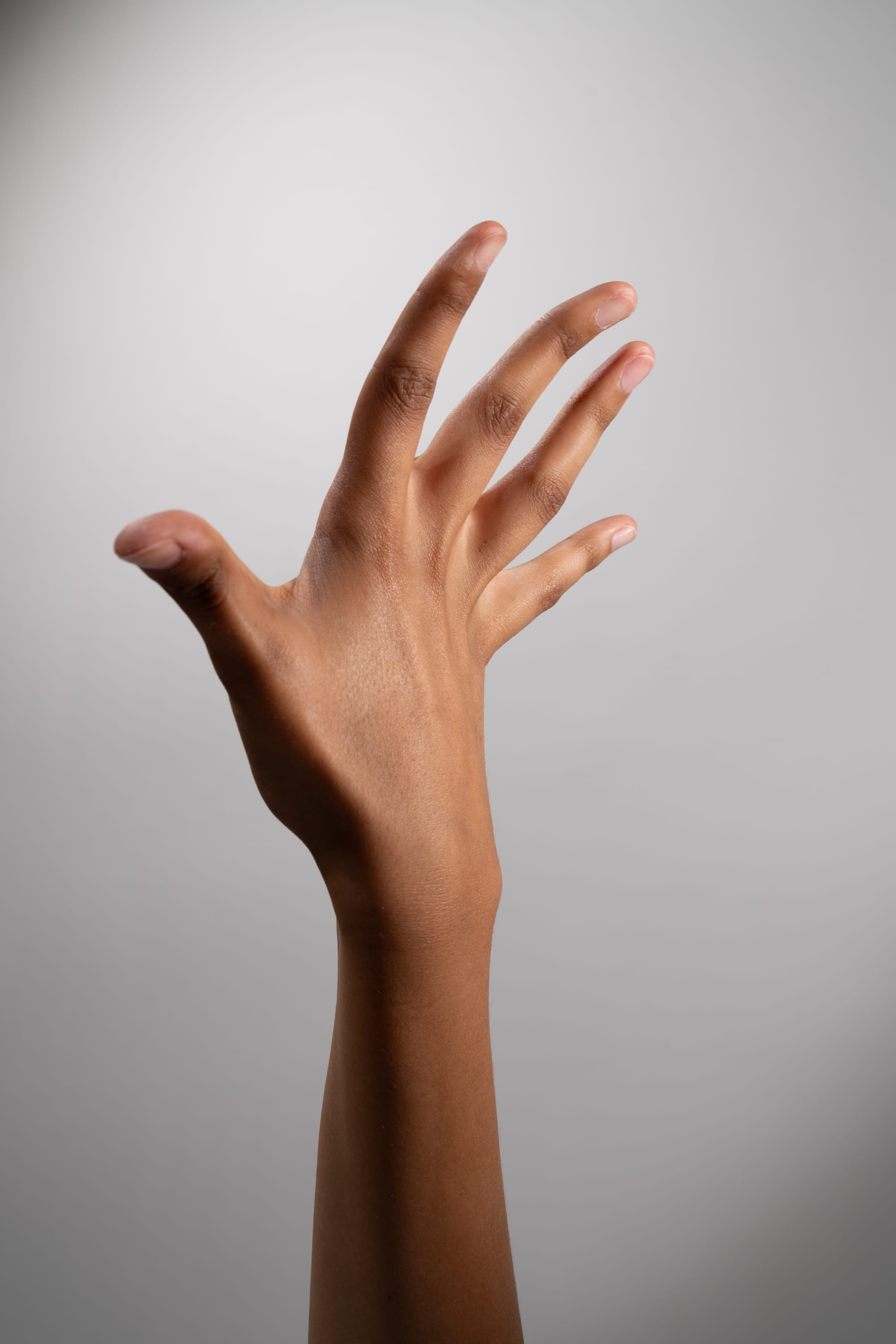 a black woman's hand is extended upward, fingers spread, against a plain grey background. the lighting is soft and even, illuminating the subtle variations in her skin tone. the hand appears to be reaching for something out of the frame.