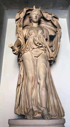 a fulllength marble statue of a roman goddess, possibly fortuna, standing on a pedestal. she holds a cornucopia in her left hand and a broken rudder in her right. she is dressed in a flowing robe that drapes over her left shoulder and arm. the statue is illuminated by natural light coming from the left side. the perspective is at eye level.