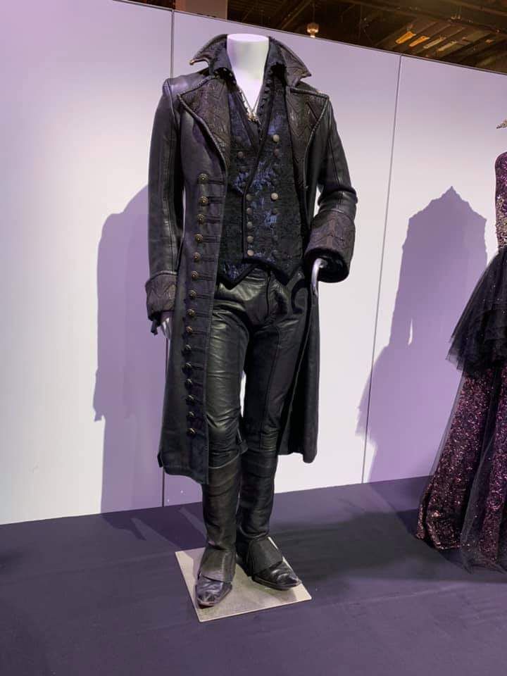 a fulllength shot showcases a mannequin dressed in a dark, elaborate costume reminiscent of a pirate or victorianera villain. the mannequin is positioned on a grey platform in front of a white panel. the costume consists of a long black leather coat with multiple buttons down the front, worn over a dark vest with intricate patterns. the mannequin also wears leather pants tucked into dark boots. the overall aesthetic is gothic and dramatic. to the right, the edge of another elaborate dress is visible.