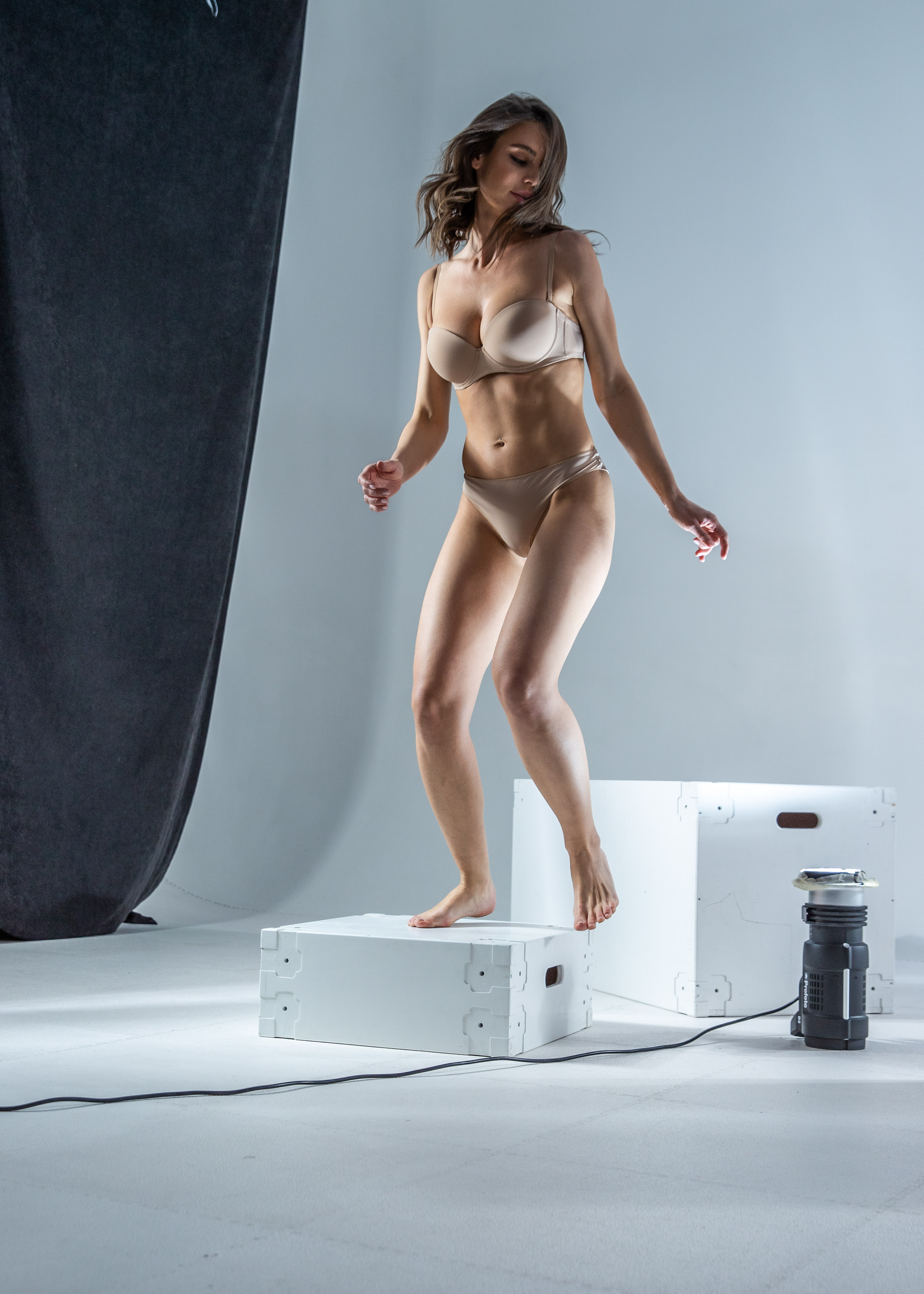 a woman wearing underwear is posing on a white box in a studio. there are other white boxes and a light nearby. there's a gray backdrop on the left side of the image. the woman is barefoot and has brown hair. she is looking down.
