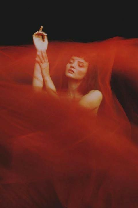 a woman is captured in a closeup shot, enveloped by a flowing, translucent red fabric. her arms are gracefully raised, and her eyes are gently closed, conveying a sense of serenity and mystery.  the red fabric dominates the frame, creating a dramatic and evocative atmosphere. the lighting is soft and moody, enhancing the overall artistic feel of the image. the woman's pose and the texture of the fabric contribute to the sensual and elegant nature of the photograph.