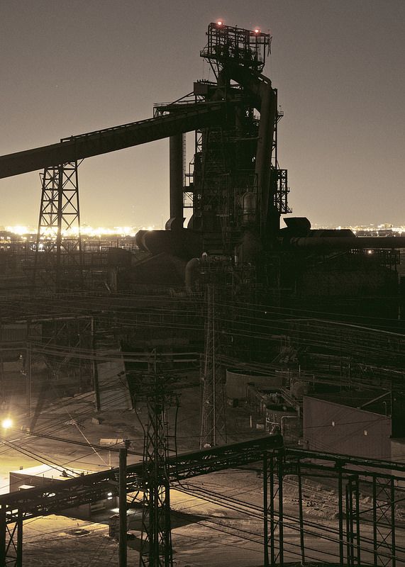 a nighttime long shot of a steel mill, possibly the bethlehem steel plant in pennsylvania. the image is dominated by the dark silhouette of massive industrial structures, including a blast furnace and extensive pipework.  the scene is lit by the internal and external lights of the plant, creating a dramatic contrast between light and dark.  the surrounding cityscape is faintly visible in the distance, further emphasizing the scale of the industrial complex.  the overall mood is one of somber grandeur, highlighting the powerful and imposing nature of heavy industry.  numerous power lines and other infrastructure crisscross the foreground and add to the complexity of the scene.