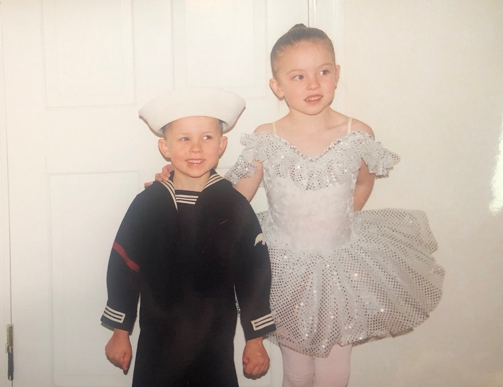 a slightly blurry vintage photograph shows a young boy and girl dressed in costume. the boy is wearing a dark blue sailor suit with a white sailor hat.  the girl is wearing a sparkly white ballet tutu. the girl has her arm around the boy's shoulders. they appear to be siblings posing for a picture indoors against a white door. the photo has a nostalgic feel, typical of older printed photos.