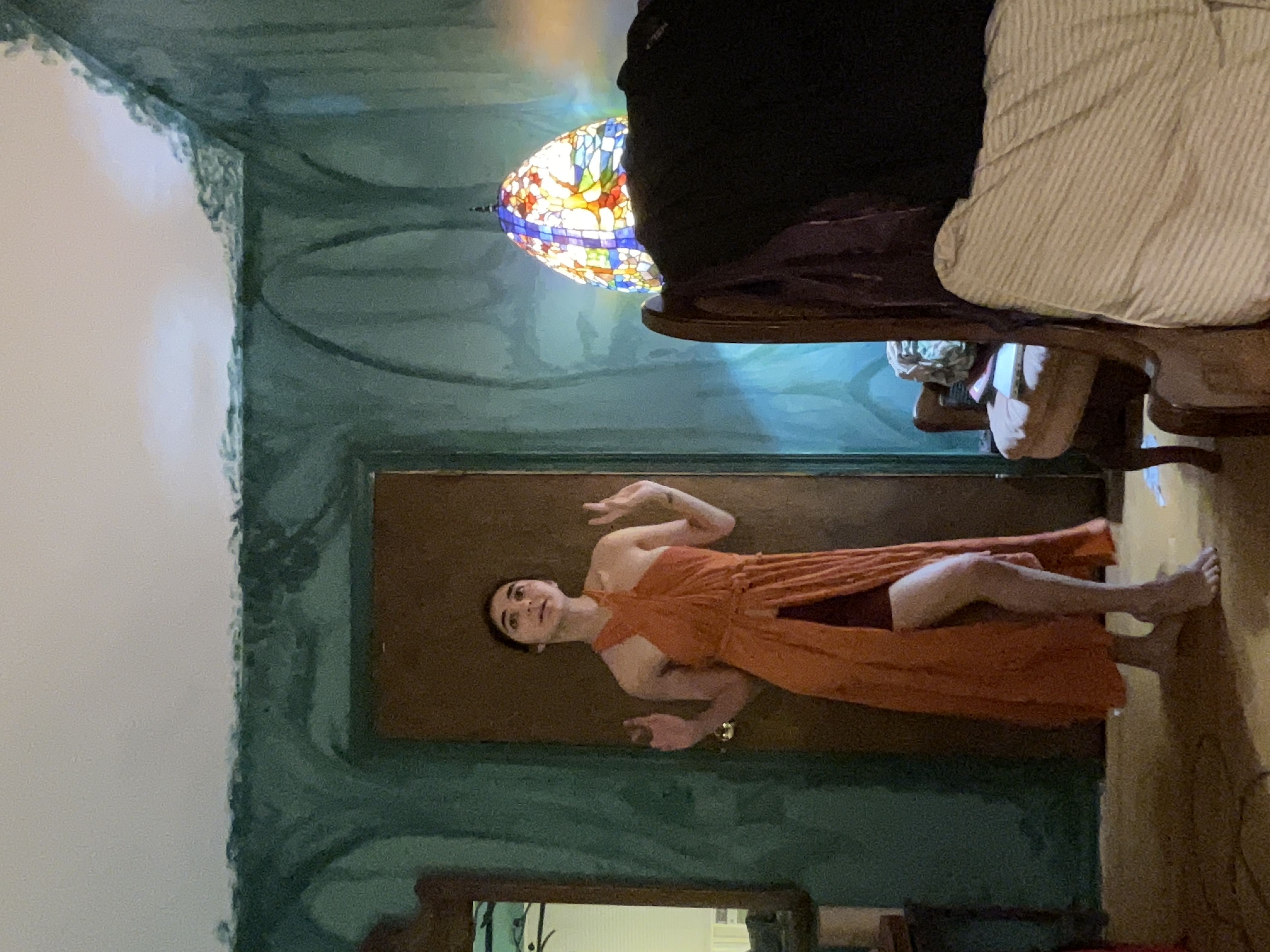 a young woman wearing a long orange halter dress with a high slit stands in a bedroom with teal walls. she is posing, her hands slightly raised. a stainedglass lamp sits on a piece of furniture in the background. the room's walls have some texture or paint imperfections.