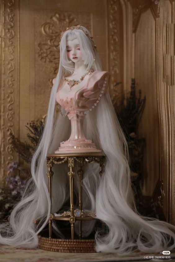 a balljointed doll with long white hair is posed on a pink and gold bust, which is placed on an ornate gold table. the doll wears a delicate necklace and the bust features a pearl embellishment. the background showcases a vintage aesthetic with floral accents and a golden framed mirror.  the overall image has a soft and dreamy quality due to the lighting and color palette.