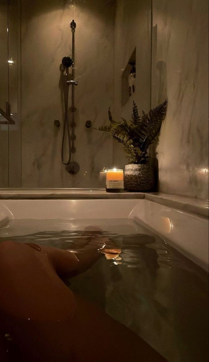 a dimly lit bathroom scene features a filled bathtub with an individual relaxing in it. the water is slightly rippled, and there's a beverage glass floating on the surface. next to the tub, there's a lit candle and a potted plant, creating a soothing ambiance. in the background, a marbletiled shower area with a showerhead can be seen, adding to the luxurious feel of the bathroom.
