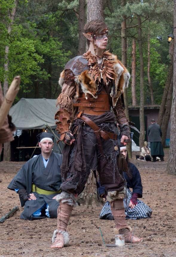 a person dressed in an elf costume stands in a forest setting, surrounded by other individuals in various costumes. the elf character wears furtrimmed clothing, leather armor, and has pointed ears, with face paint. the background shows trees, a tent, and other people in costume. the overall scene suggests a liveaction roleplaying event or a cosplay gathering in a natural environment.