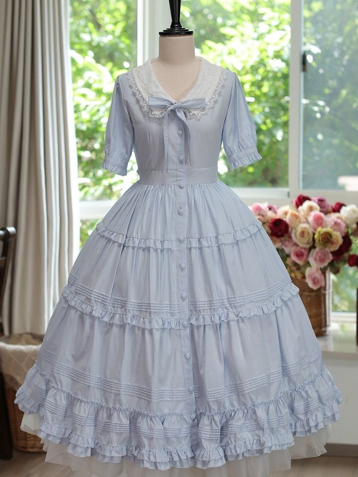 a light blue lolita fashion dress is displayed on a mannequin in front of a window. the dress features short puff sleeves with white lace trim, a white lace collar with a bow tie, a buttonup front, and a tiered skirt with ruffles. the dress is long and flowy, reaching the floor. there is a window behind the mannequin with soft, natural light streaming in. a vase of flowers is visible to the right of the mannequin.