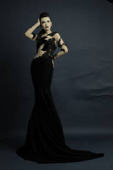 a fulllength studio shot of a woman model wearing a stunning black mermaidstyle gown. the dress is made of sheer fabric adorned with intricate black lace appliqués, creating a seethrough effect that showcases her figure while maintaining elegance. the dress flows gracefully to the floor, creating a dramatic silhouette.  her hair is styled up, and her makeup is sophisticated and enhances her features. the background is a dark gray, which contrasts beautifully with the black dress, making the model the clear focal point of the image. the overall mood is one of high fashion and glamour.