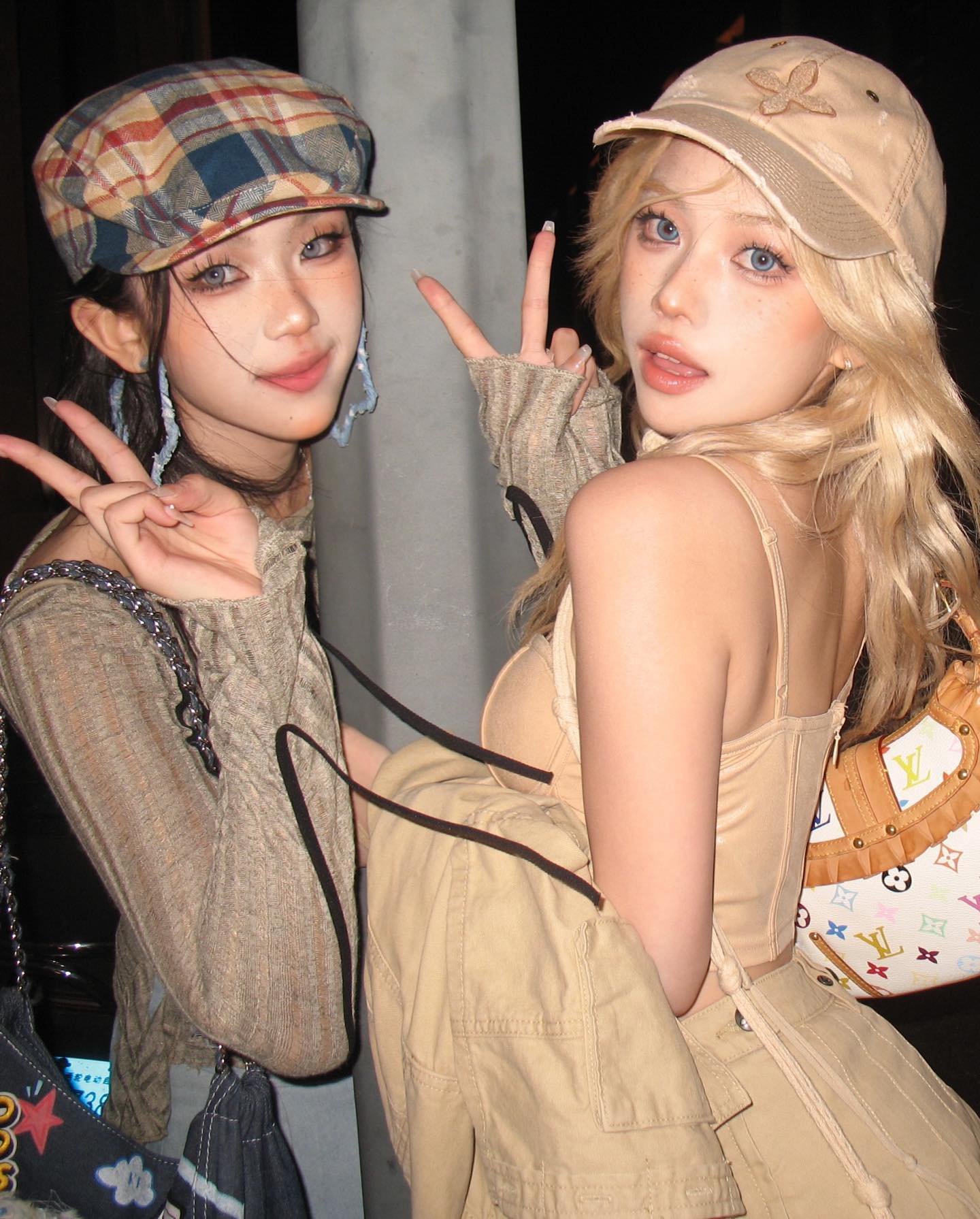 two young women pose for a selfie on a city street at night. the woman on the left wears a brown plaid newsboy cap, a brown patterned top, and large blue earrings. the woman on the right wears a beige baseball cap, a beige corset top, and beige cargo pants. both women are holding up peace signs. the photo has a vintage, y2k aesthetic.