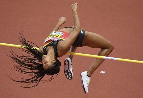 a female athlete leaps over a high jump bar with her back arched and her body in midair. she is focused on clearing the bar, her face contorted in concentration. her long hair flows behind her, blurred by the speed of her movement. the image captures the athleticism and grace of a high jumper in action.