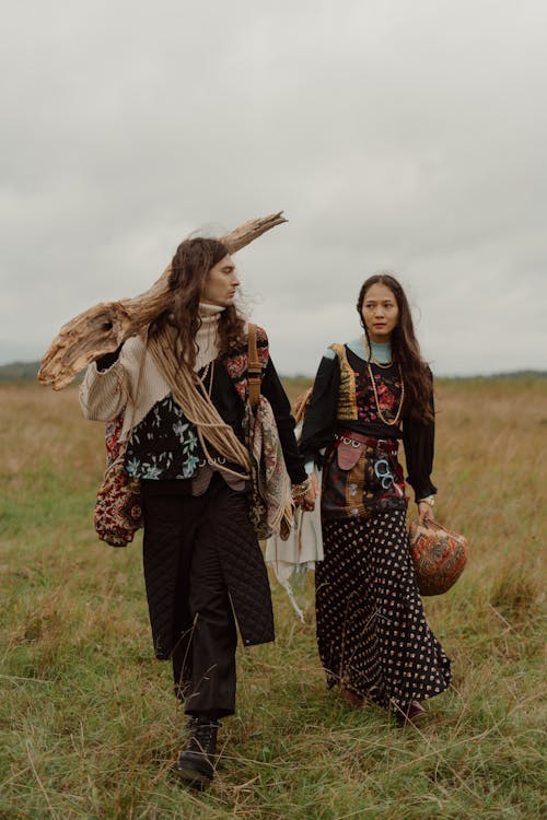 a man and a woman, dressed in eclectic bohemianstyle clothing, walk handinhand across a grassy field. the man carries a large piece of driftwood across his shoulders, and both individuals wear layered clothing with patchwork and textured fabrics. the overall atmosphere is vintage and retro, with a focus on unique fashion choices and natural surroundings. the scene is set in an outdoor setting in autumn or fall.