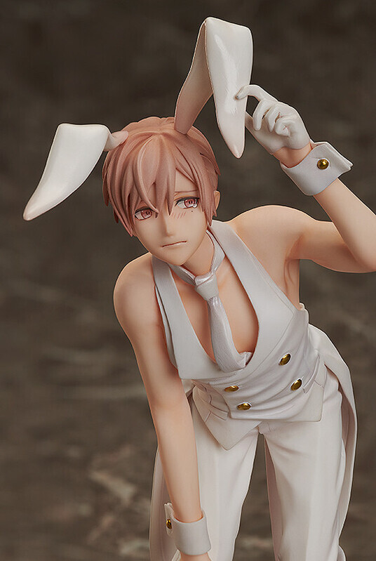 loseup view of an anime figurine depicting a young man with pink hair dressed in a white bunny suit.  he's wearing a white tuxedostyle outfit with a waistcoat, and he has bunny ears atop his head. his expression is shy or embarrassed, with a slight blush on his cheeks. the figurine is highly detailed, showing a smooth texture and realistic features. the background is blurred, focusing the viewer's attention on the figure.  the pose suggests a slightly hunched and demure stance.