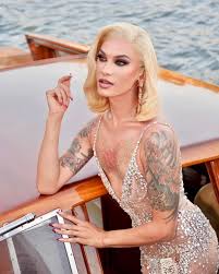 a glamorous drag queen with blonde hair and tattoos poses on a luxurious boat on the water. she is wearing a sparkling, sheer, lowcut gown and looks directly at the camera. the scene is set against a backdrop of water. the image conveys luxury and elegance.