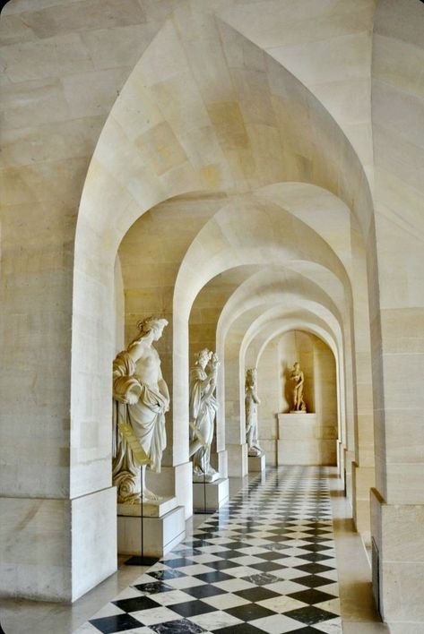 a long corridor with high vaulted ceilings is lined with white marble statues on pedestals. the floor is a checkered pattern of black and white tiles. the corridor features a series of arches that create a sense of depth and grandeur. the overall impression is one of elegance and sophistication.