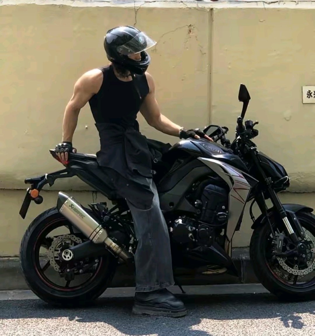 a muscular man in a black sleeveless shirt and grey pants is sitting on a black kawasaki z900 motorcycle. he's wearing a black helmet and gloves. the motorcycle is parked against a light beige wall. the scene is set outdoors on a sunny day.  the image suggests a sense of style and power.