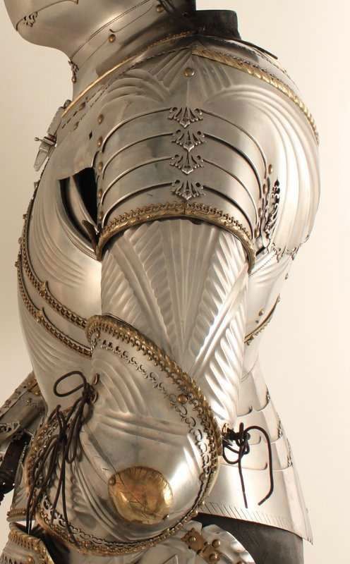 this is a closeup image showcasing the intricate detail of a suit of armor.  the armor is made of polished metal with a series of etched lines that create a fluted effect on the breastplate and pauldrons. decorative gold accents are visible on the edges of the armor pieces. the armor is laced together with leather straps, suggesting it is flexible and designed for combat.  the background is white or offwhite, putting the focus solely on the craftsmanship of the armor.