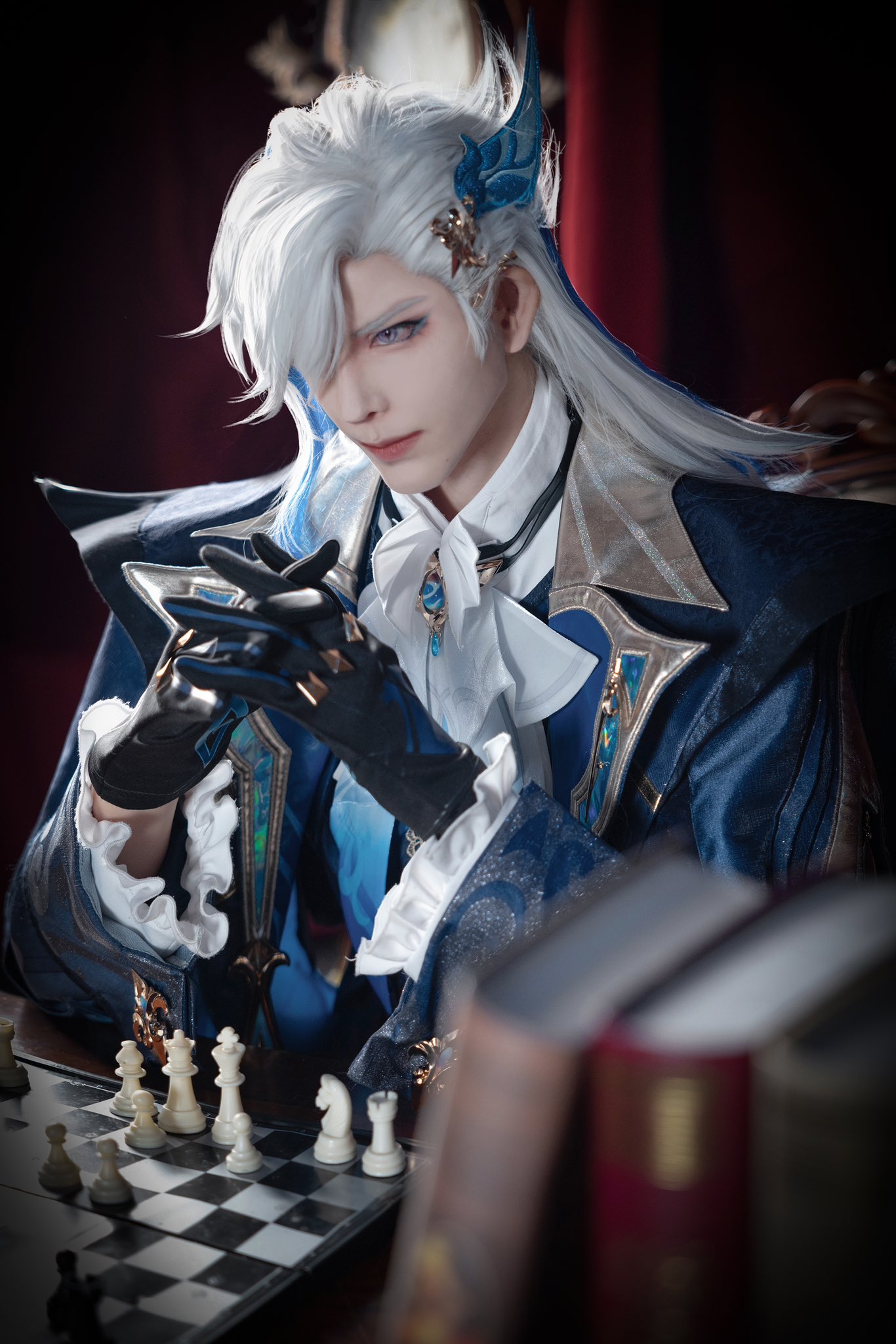 a young man in an anime cosplay, with white hair and blue eyes, is wearing a blue and gold coat. he is wearing black gloves and is sitting at a chessboard, with his hands clasped together. the chessboard is in the foreground and there are books on the table behind him. the image is set in a dark, shadowy room, with the only light coming from the windows behind the man.
