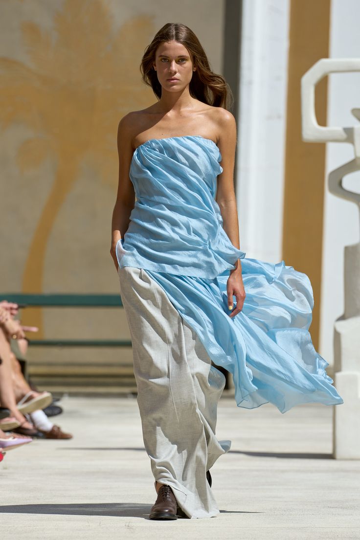 a young woman is posing on a runway. she is wearing a blue dress with white pants and sandals. her hair is flowing in the wind. she is looking towards the camera.
