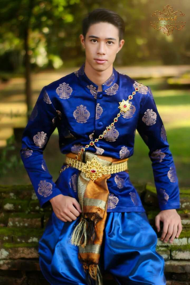 a young thai man is wearing traditional clothing. he is smiling and looking at the camera.  his clothing is a blue shirt with gold embroidery and a gold sash around his waist.  the sash is tied with a large gold ornate knot in the front.