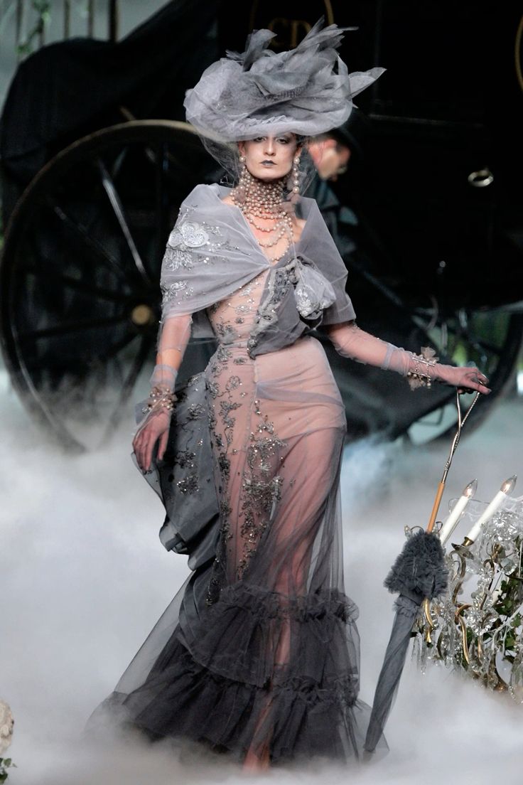 a fashion model walks the runway in a stunning gray and silver gown with intricate lace detailing. her look is both elegant and whimsical, featuring a sheer tulle skirt, a delicate lace bodice, and a large, dramatic hat. the model's hair is styled in a loose, tousled wave, and her makeup is subtle and ethereal. the overall effect is one of sophisticated glamour and ethereal beauty. the model is adorned with delicate silver jewelry that complements her outfit perfectly. her pose is confident and captivating, and she carries herself with an air of grace and poise. this image captures the essence of high fashion, showcasing the artistry and craftsmanship of the designers. the setting is a grand and opulent, creating a sense of fantasy and enchantment. the model's outfit and the overall ambiance of the scene evoke a sense of timeless elegance and beauty.