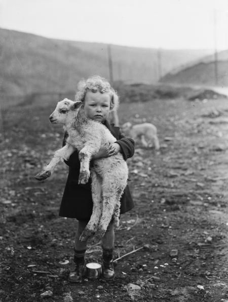 a young girl in a black coat holds a small white lamb in her arms. she is standing outdoors, likely on a farm or in the countryside. the photo is in black and white.