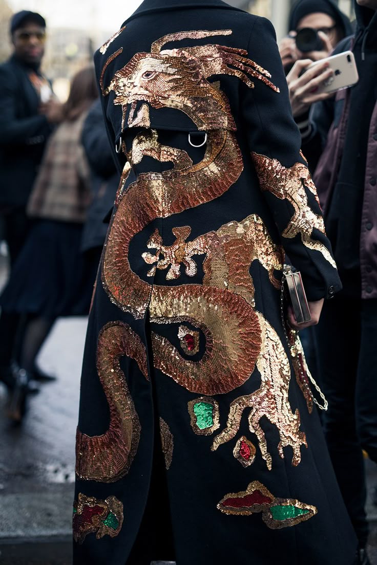 a black coat with a large golden dragon embroidered on the back is shown in this image. the dragon is embellished with golden sequins, and has red and green gemstones along its body. the coat is being worn by a person standing on a street, surrounded by other people.