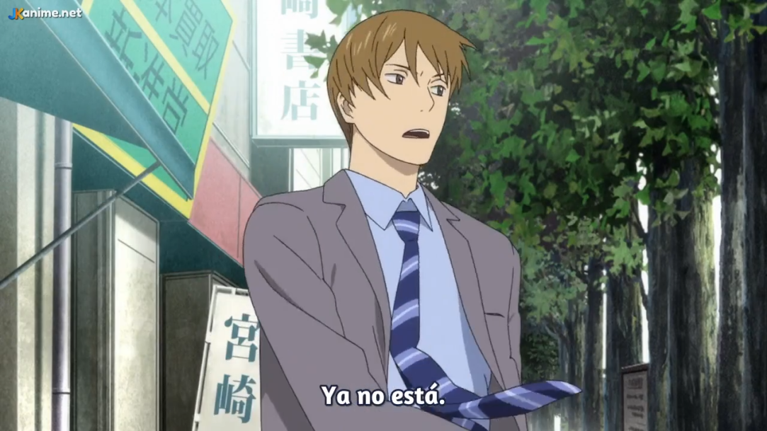 an anime still frame featuring a young man in a light grey suit and blue striped tie standing on a city sidewalk. he appears surprised or concerned, with his mouth slightly open and eyebrows raised. he's holding his tie loosely with one hand. the background depicts a street scene with buildings, trees, and signage written in japanese. the overall lighting is soft and diffused, suggesting an overcast daytime setting.
