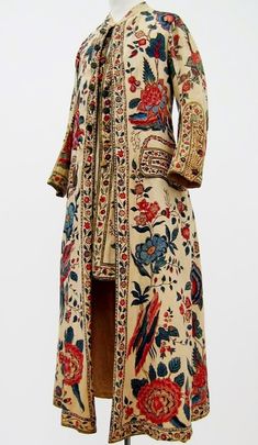 a vintage floral dress with long sleeves.  the floral print is red, blue, and white.  the dress has a cream background, with a trim around the neckline and a trim down the front. the dress is detailed and ornate.