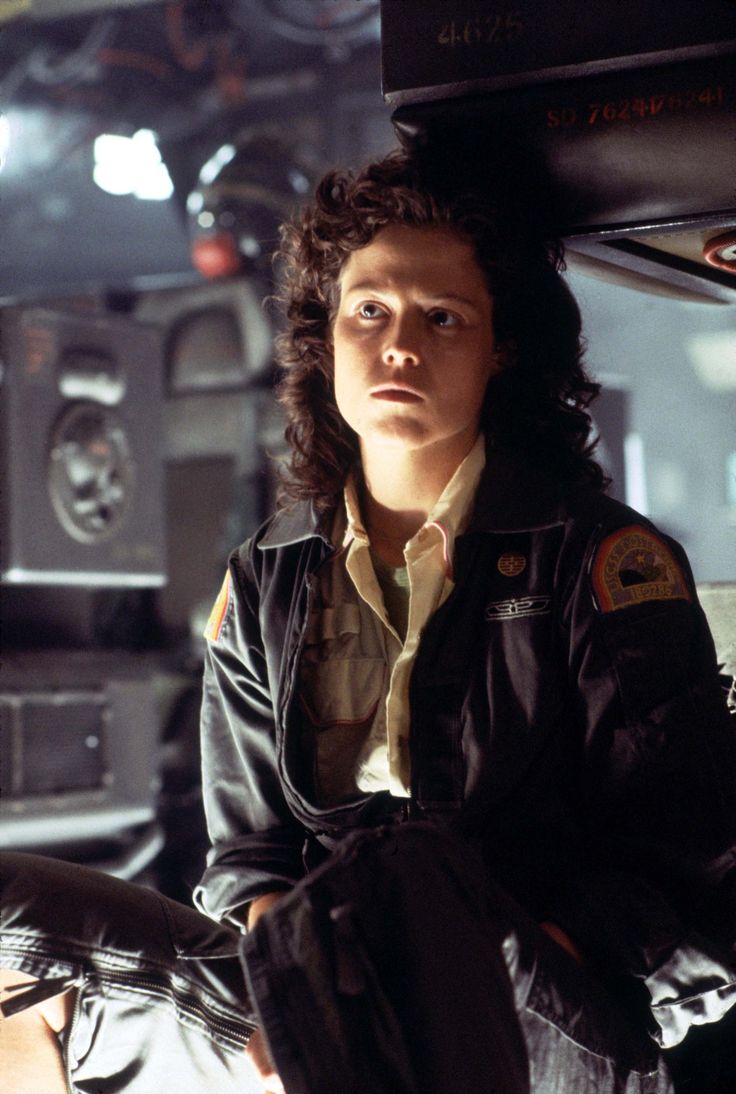 this is a still from the movie "alien", where the actress, sigourney weaver who plays ellen ripley, looks up with a serious expression on her face, looking concerned as she sits in the interior of the spaceship with the crew.