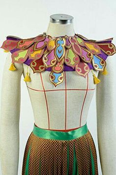 this is an image of a princess jasmine inspired costume piece, a colorful bejeweled collar or bibstyle necklace.  the base of the neckpiece appears to be gold and it is decorated with stylized "petals" or "leaves" in a variety of colors and patterns, with yellow tassels hanging from the two bottom points. the mannequin is also wearing a green waistband over a brown skirt, possibly part of a costume.  this image appears to have been taken indoors.
