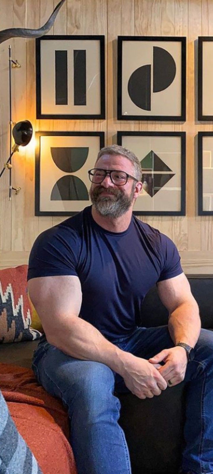 a muscular man with a beard and glasses sits on a couch, wearing a navy tshirt and jeans. there is wall art in the background.